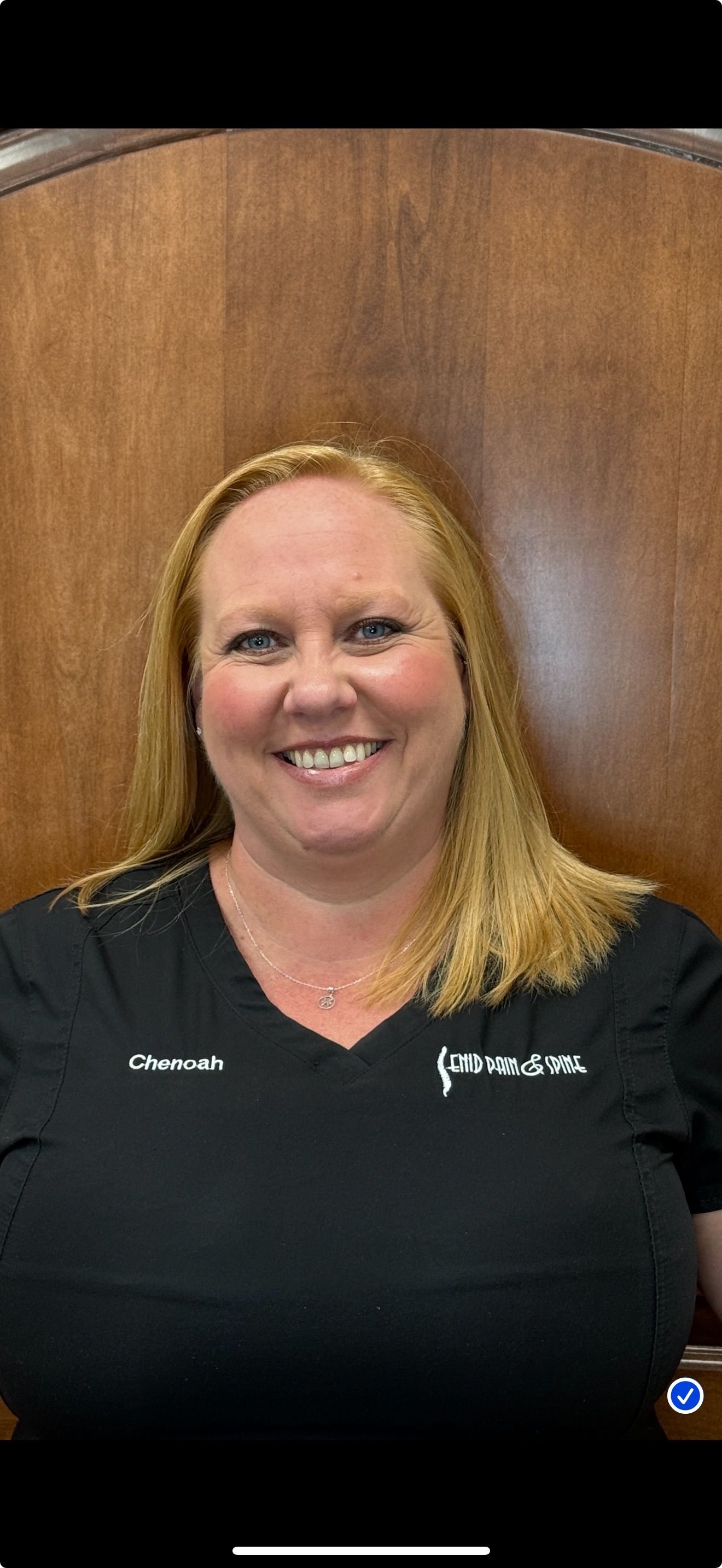 Woman in black scrubs smiles in front of a wood background.