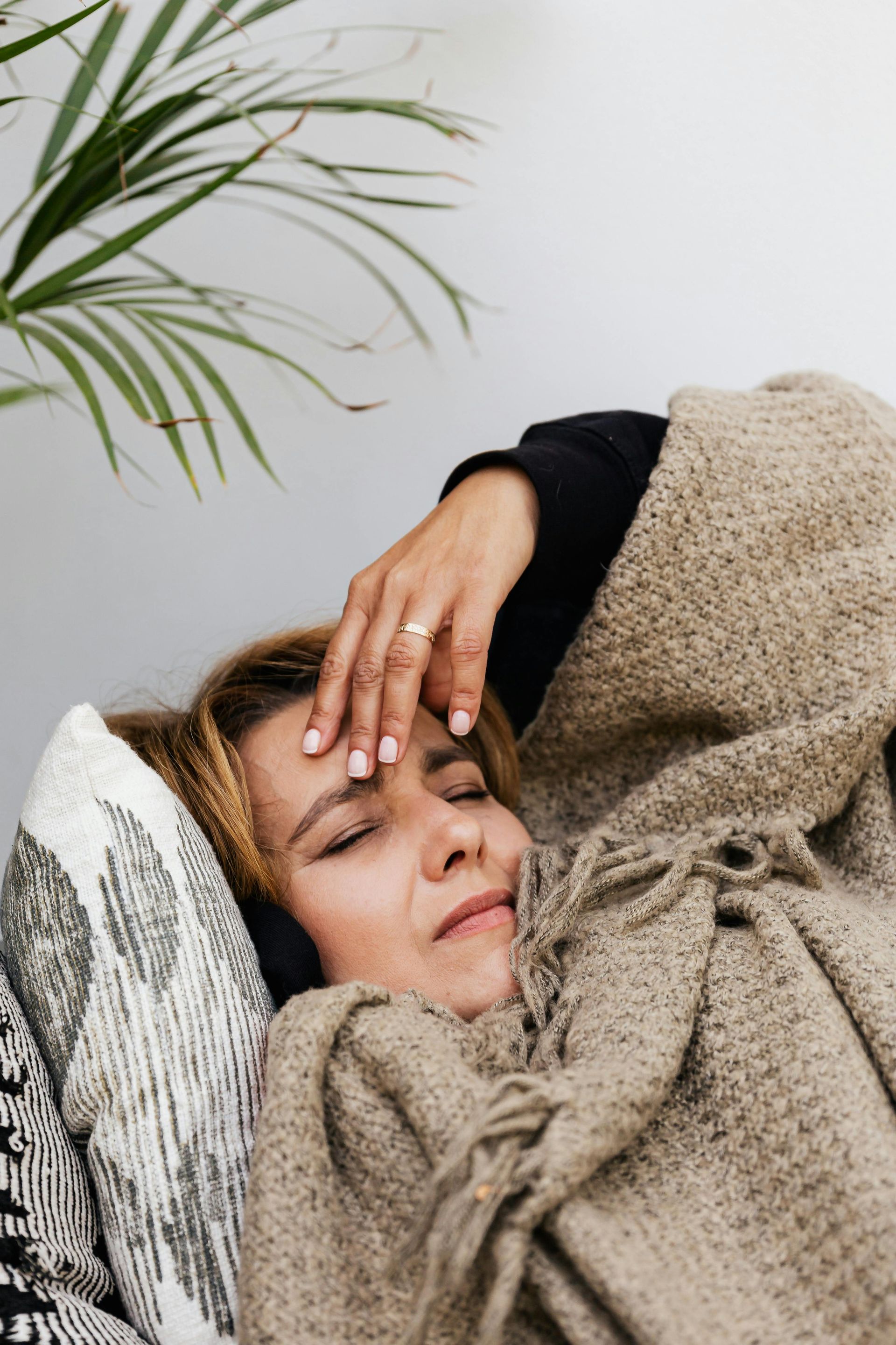Woman lying down, touching forehead, wrapped in a blanket, near a plant; appears unwell.