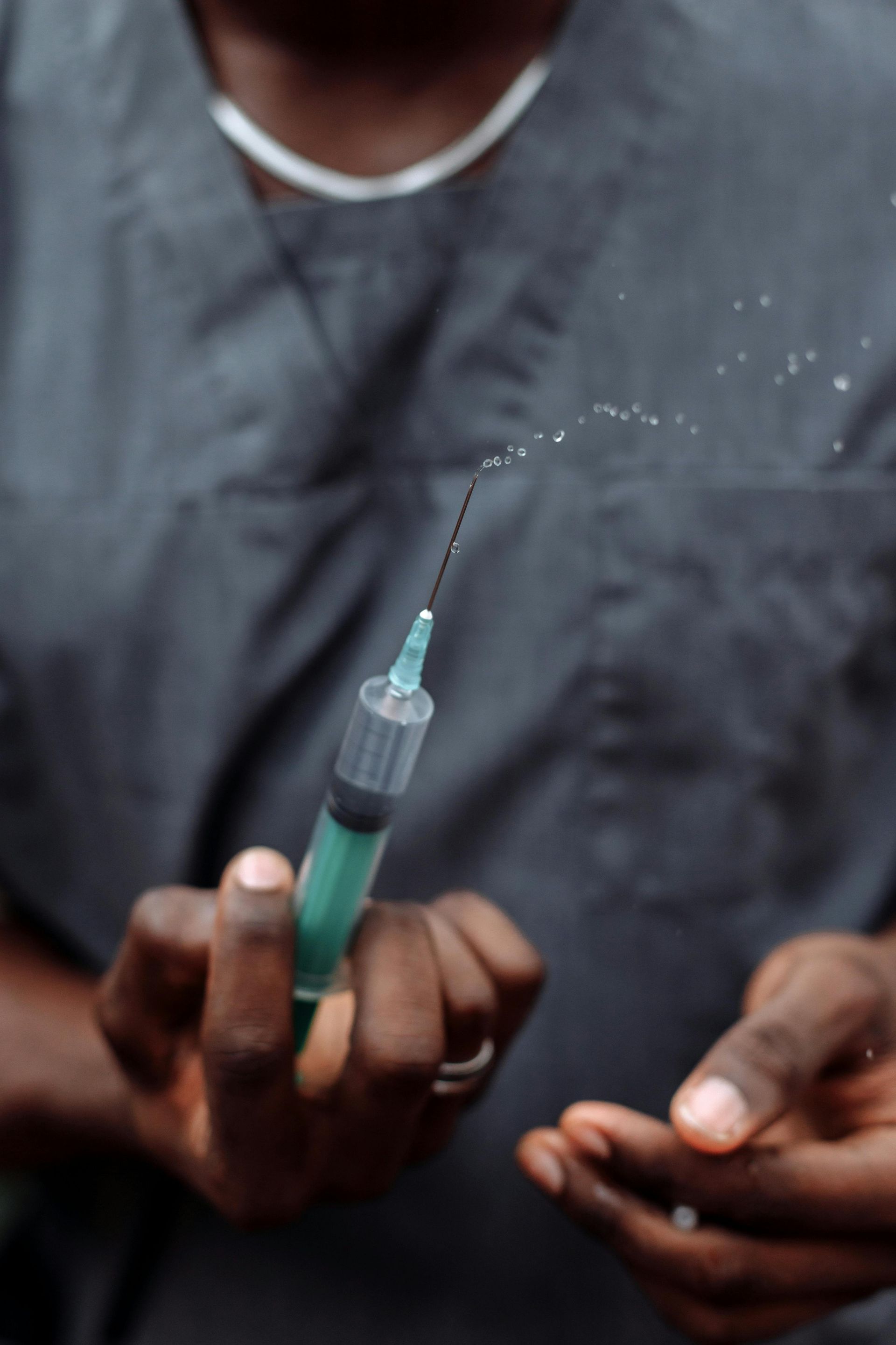Hands holding a syringe, squirting liquid. The person wears grey scrubs.