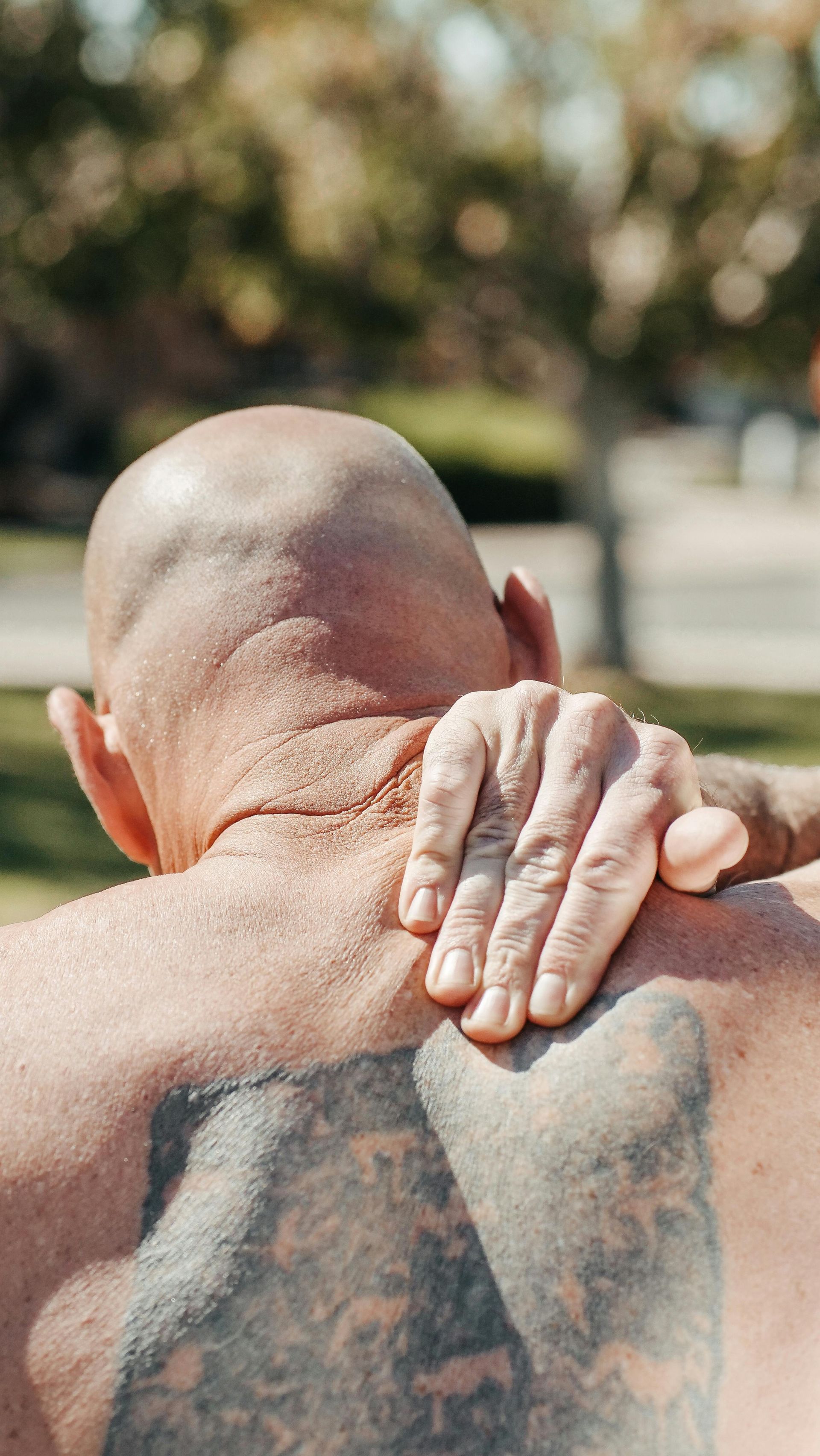 Bald person with tattoo on back touches shoulder outdoors.