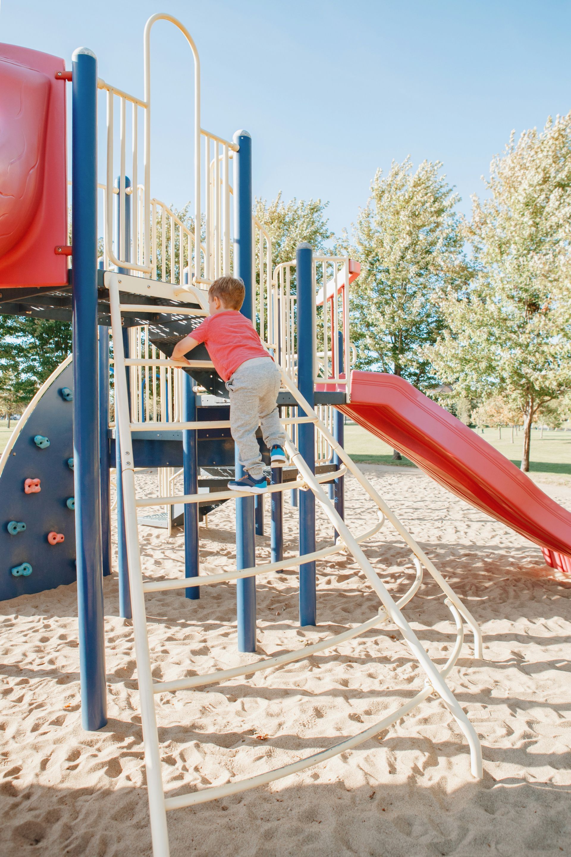 Young child climbing a metal ladder on a playground, sandy ground, sunny day.