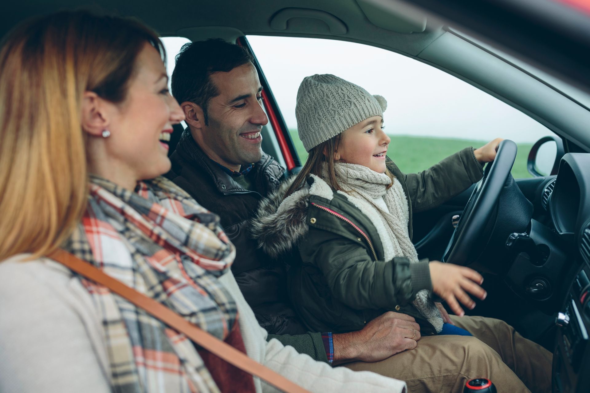 Family in car; child pretends to drive, parents smile, green field visible.