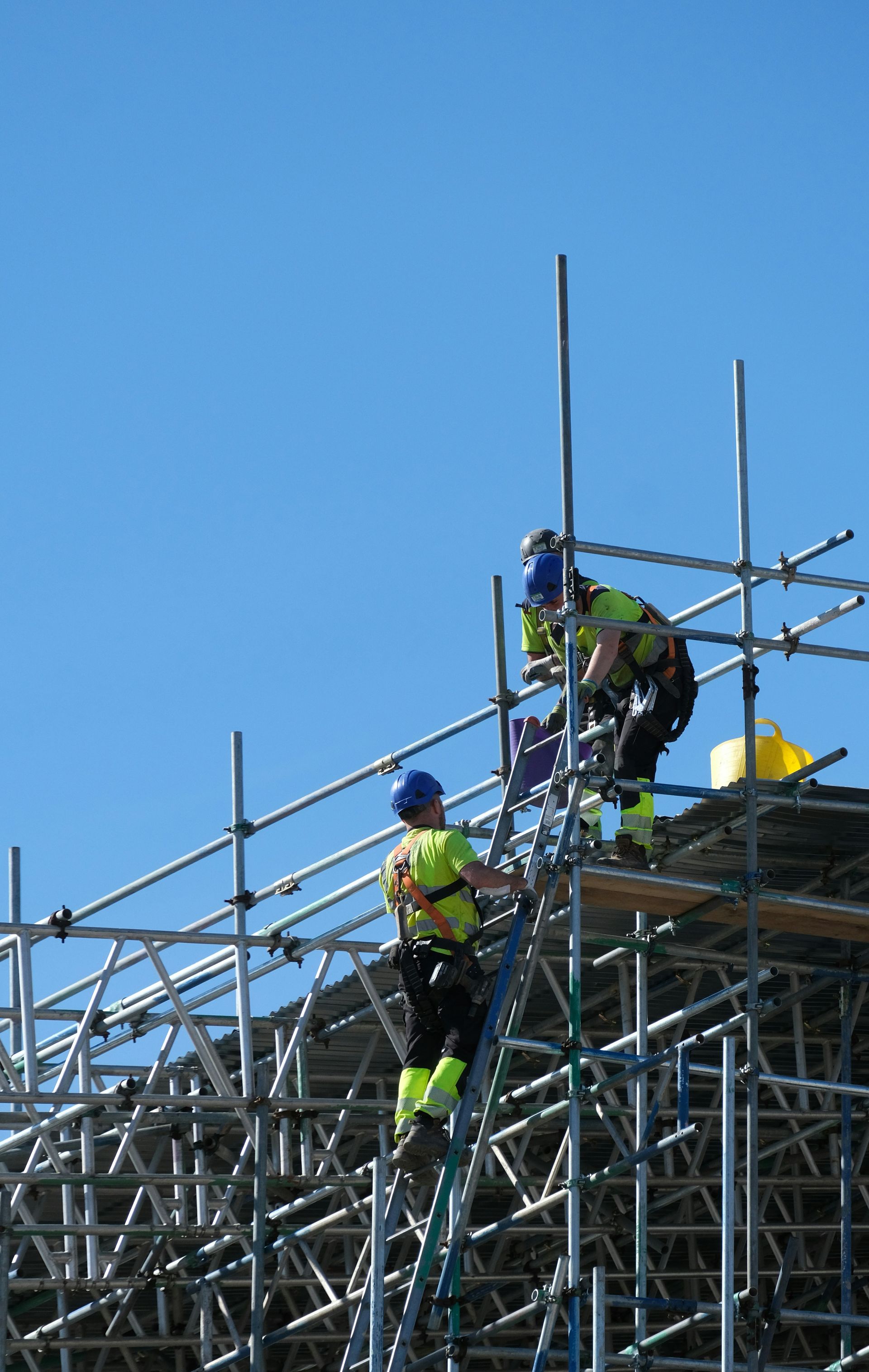 Construction workers on scaffolding, wearing safety gear, working on a sunny day.