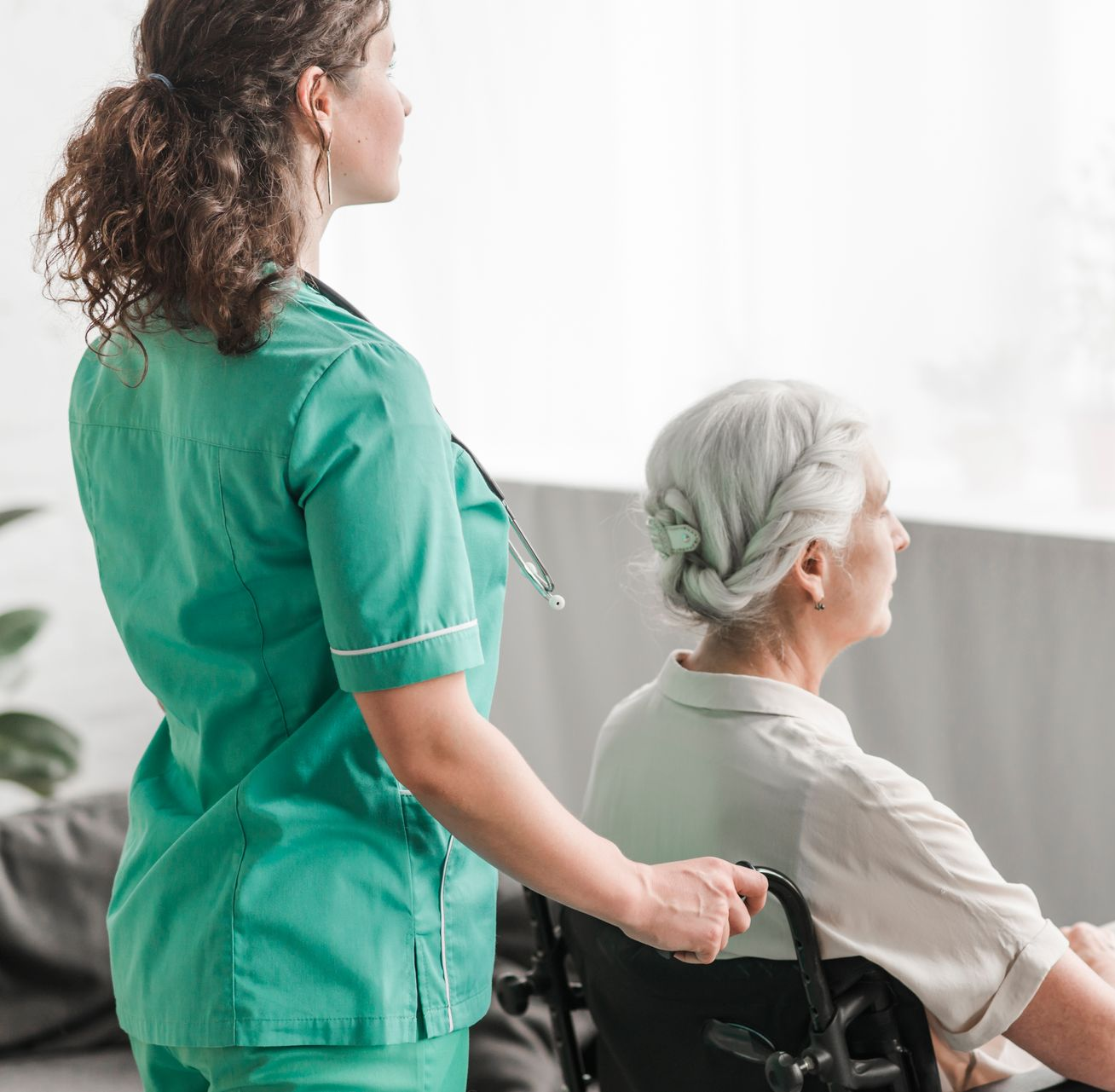 Nurse in green scrubs pushing a person in a wheelchair indoors near a window.