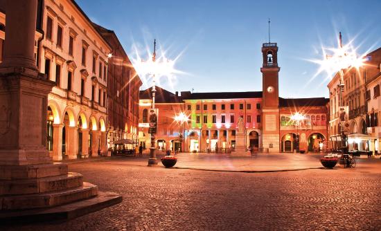 Piazza acciottolata al tramonto, circondata da edifici con archi e una torre dell'orologio. Luci intense si riflettono.