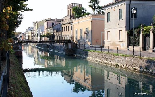 Canale in una città europea con edifici che costeggiano le rive; ponte; l'acqua riflette gli edifici e il cielo.