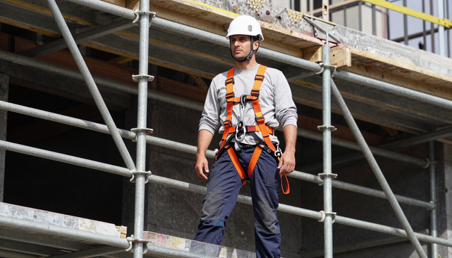 Operaio edile con imbracatura di sicurezza e casco protettivo in piedi su un'impalcatura in un cantiere.