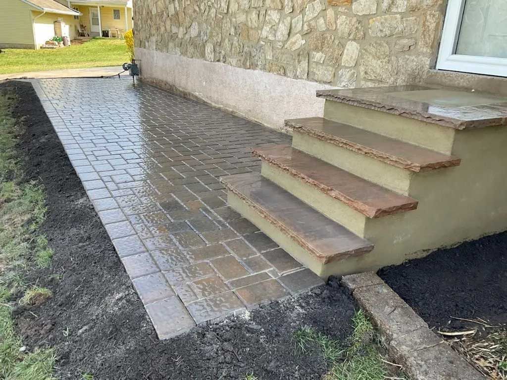 A stone patio leads to three steps with brown stone treads at the base of a textured stone house wall with dark mulch. A stone patio leads to three steps with brown stone treads at the base of a textured stone house wall with dark mulch.