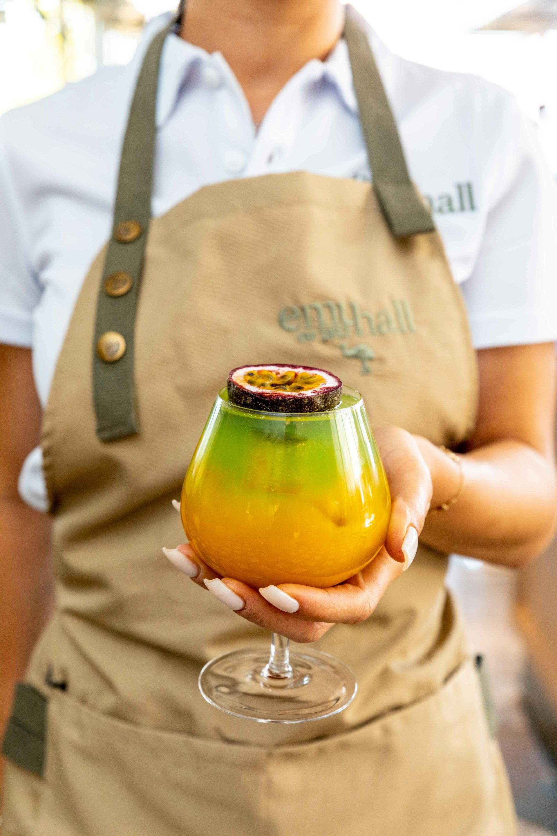 Woman holding a layered cocktail with passion fruit garnish; wearing a white shirt and tan apron.