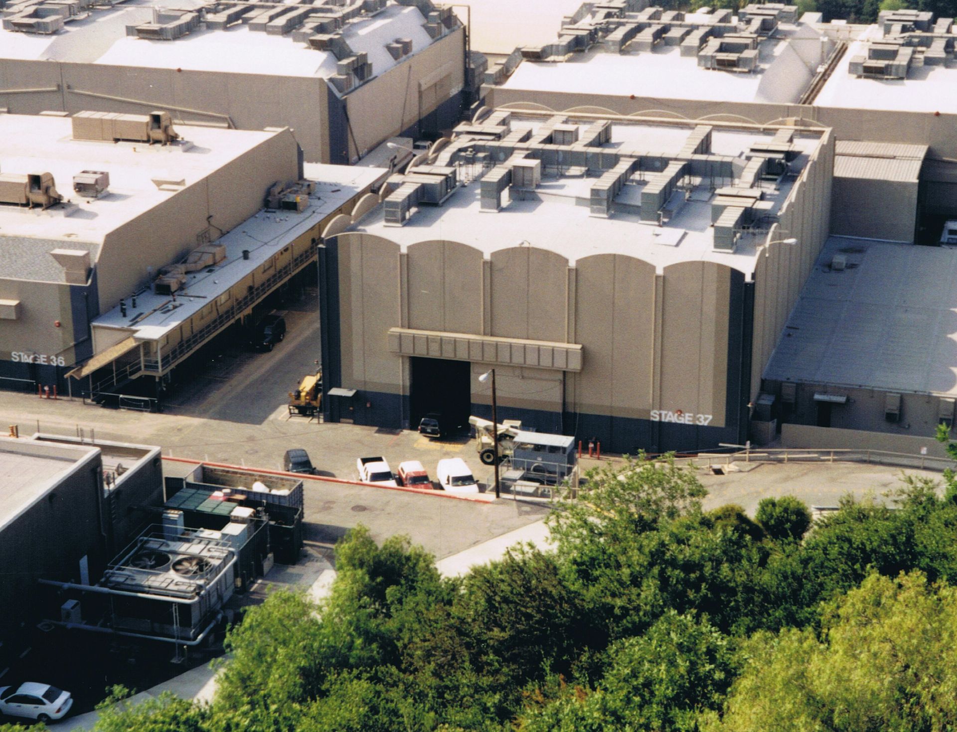 Exterior view of large beige movie studio buildings with cars and greenery nearby.