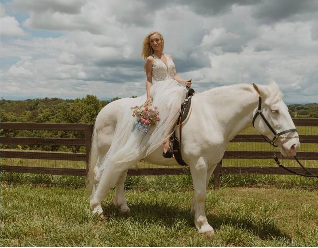 Bride in a white dress, riding a white horse in a field; holding flowers.
