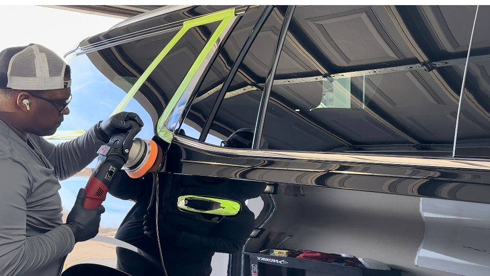 Man polishing black car door in a garage with a rotary polisher, wearing gloves, cap, and a long-sleeve shirt.