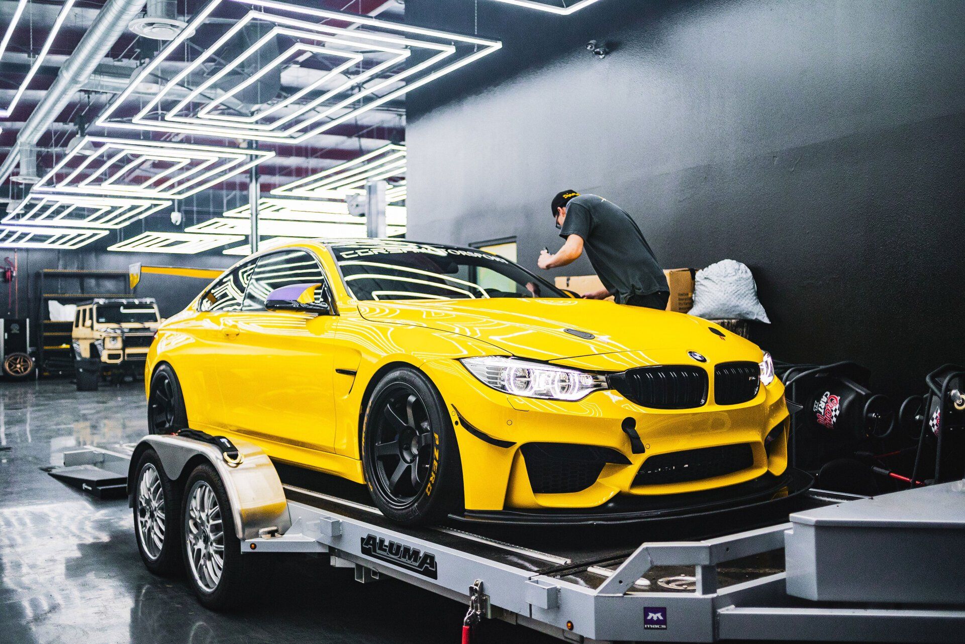 Yellow BMW M4 on a trailer being worked on inside a shop. Black wheels and a man in black shirt.