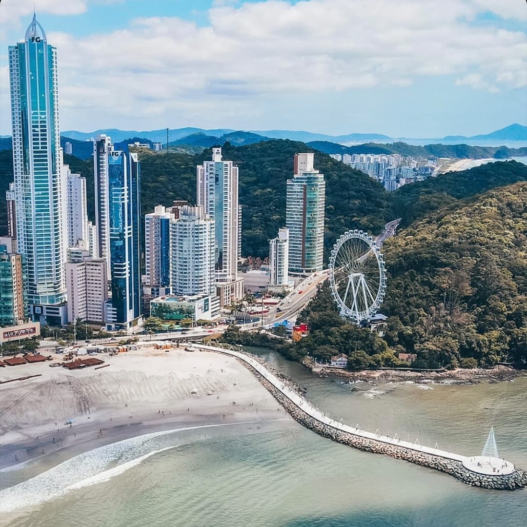 Cidade costeira com arranha-céus, praia, píer e roda-gigante. Colinas verdejantes ao fundo. Céu azul e oceano.