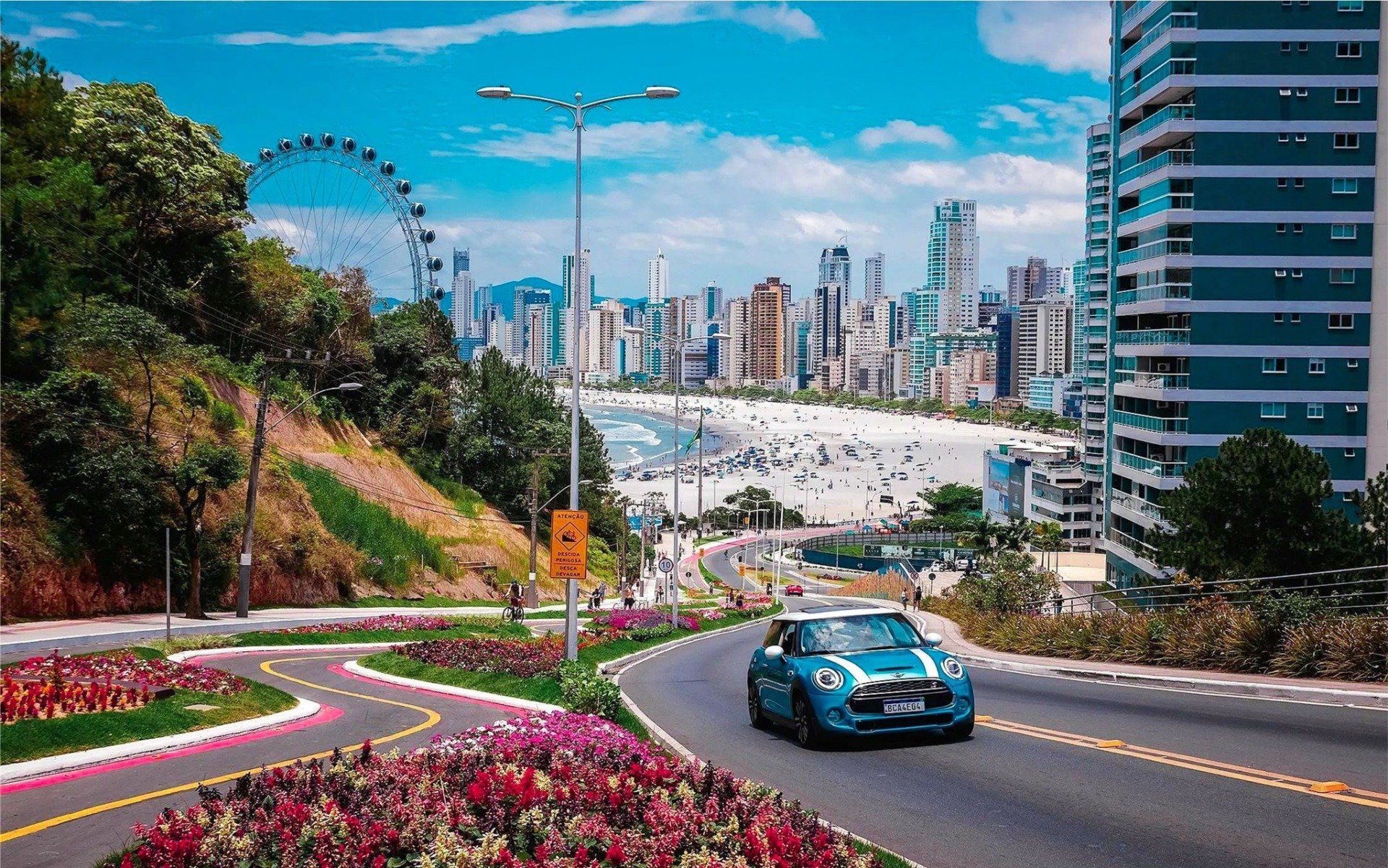 Carro azul dirigindo em uma estrada sinuosa, com uma praia, paisagem urbana e roda gigante ao fundo.
