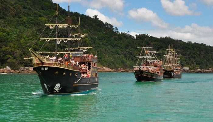 Três navios piratas em águas azul-turquesa, navegando perto de um litoral verde e montanhoso.
