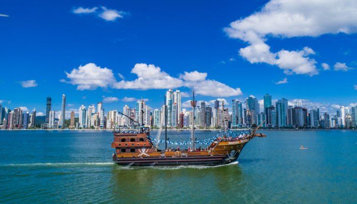 Navio pirata navegando em águas azuis, com o horizonte da cidade ao fundo, sob um céu azul.