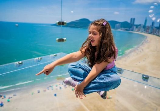 Jovem aponta para baixo, sentada em uma plataforma de vidro com vista para a praia e o oceano.