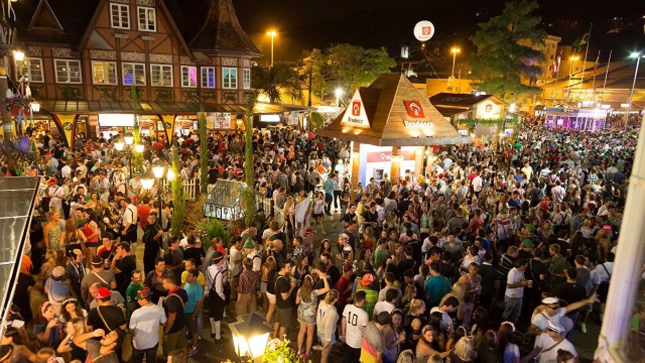 Grande multidão em uma praça festiva à noite; prédios com luzes; pessoas socializando.