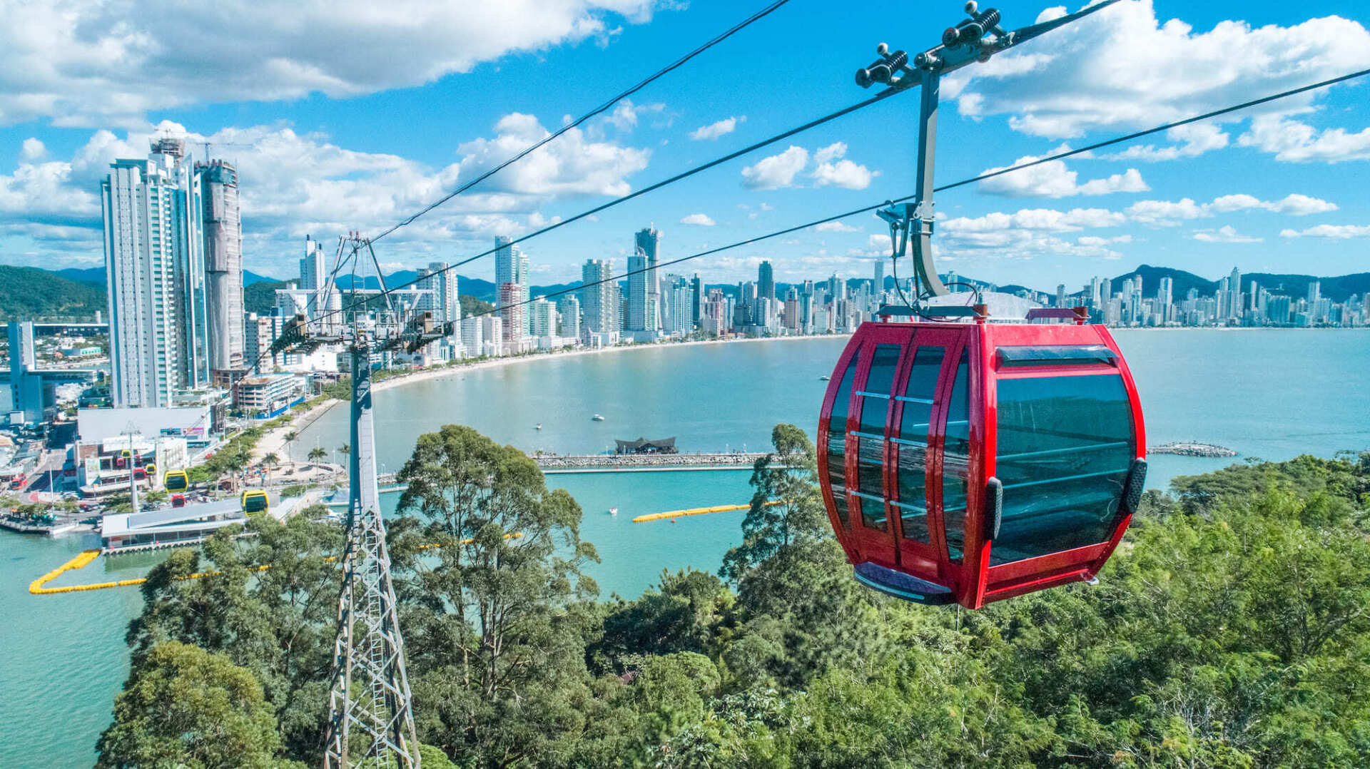 Teleférico vermelho contra o horizonte de uma cidade costeira, céu azul, folhagem verde.