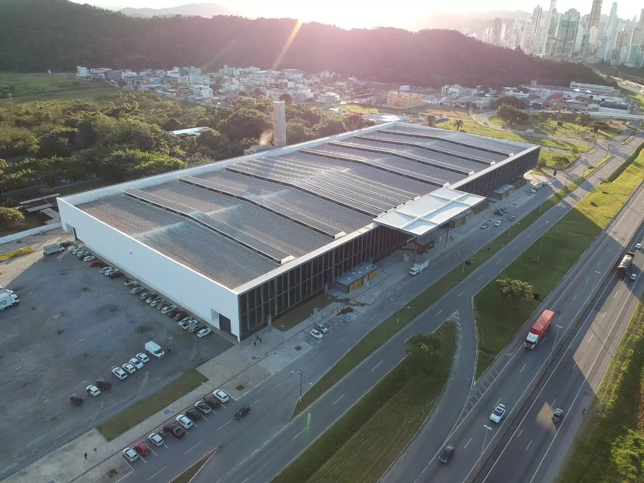 Vista aérea de um grande armazém com estacionamento e uma rodovia, em frente a uma cidade e montanhas.