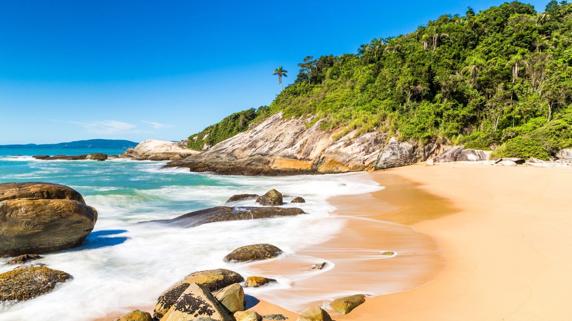 Praia de areia com águas azul-turquesa, pedras e uma colina verdejante sob um céu azul brilhante.