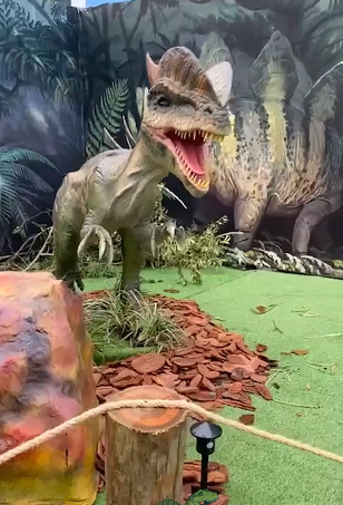 Um modelo de dinossauro com a boca aberta em uma exposição temática, com um estegossauro ao fundo.