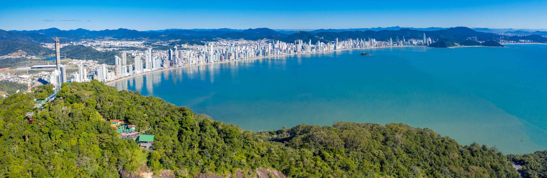 Vista aérea de uma cidade costeira com muitos prédios altos, ao lado de um grande corpo de água azul-turquesa. Colinas verdes em primeiro plano.