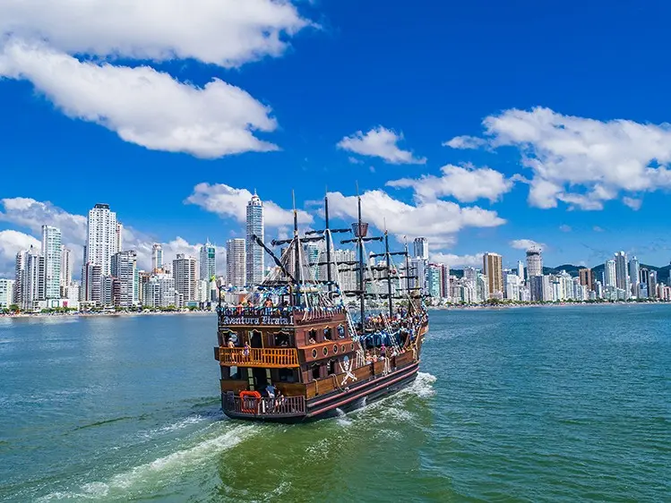 Navio pirata navegando em águas azuis, com o horizonte da cidade ao fundo, sob um céu azul com nuvens brancas.