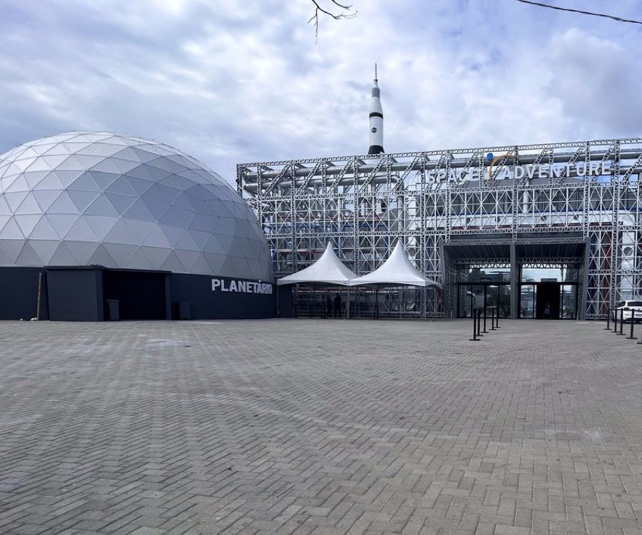 Exterior de um edifício com tema espacial, com uma cúpula de planetário, estrutura de metal e entrada com um foguete.
