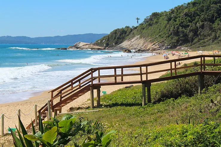 Uma passarela de madeira leva a uma praia de areia com ondas; uma encosta verde ao fundo sob um céu ensolarado.