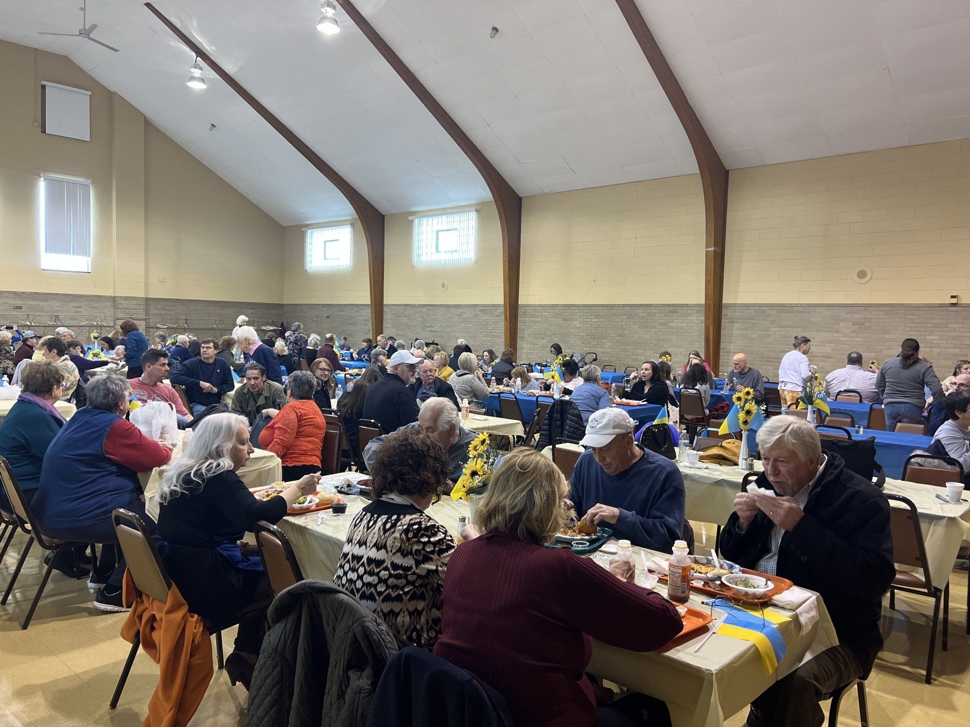 Large community hall with many people seated at tables, eating and socializing during a gathering