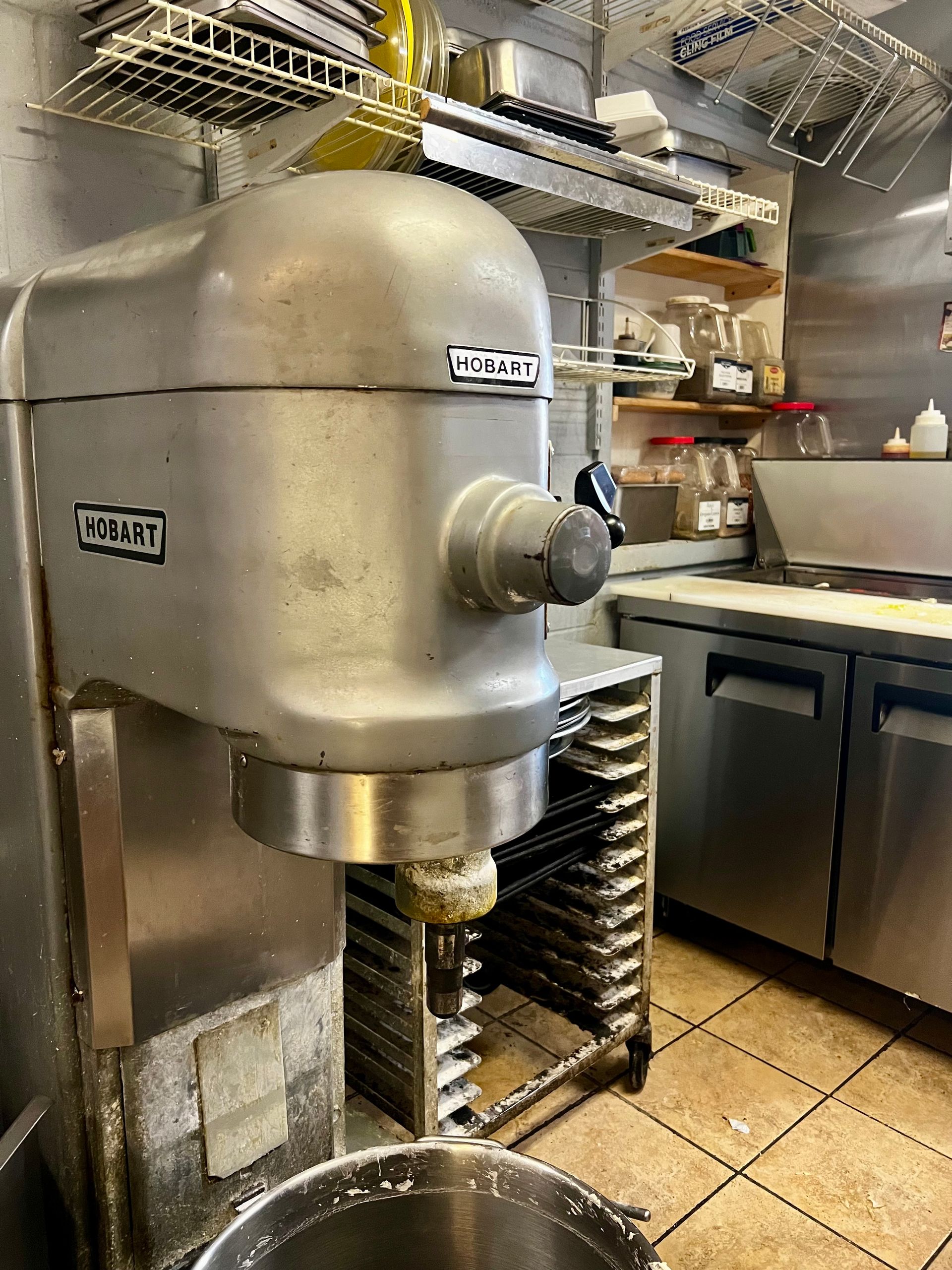 Large silver commercial mixer in a stainless-steel kitchen, with a rack of trays beneath it.