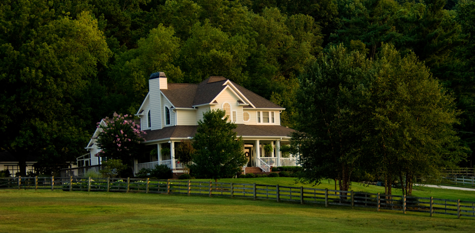 White farmhouse with porch in a green field, surrounded by trees and a wooden fence.