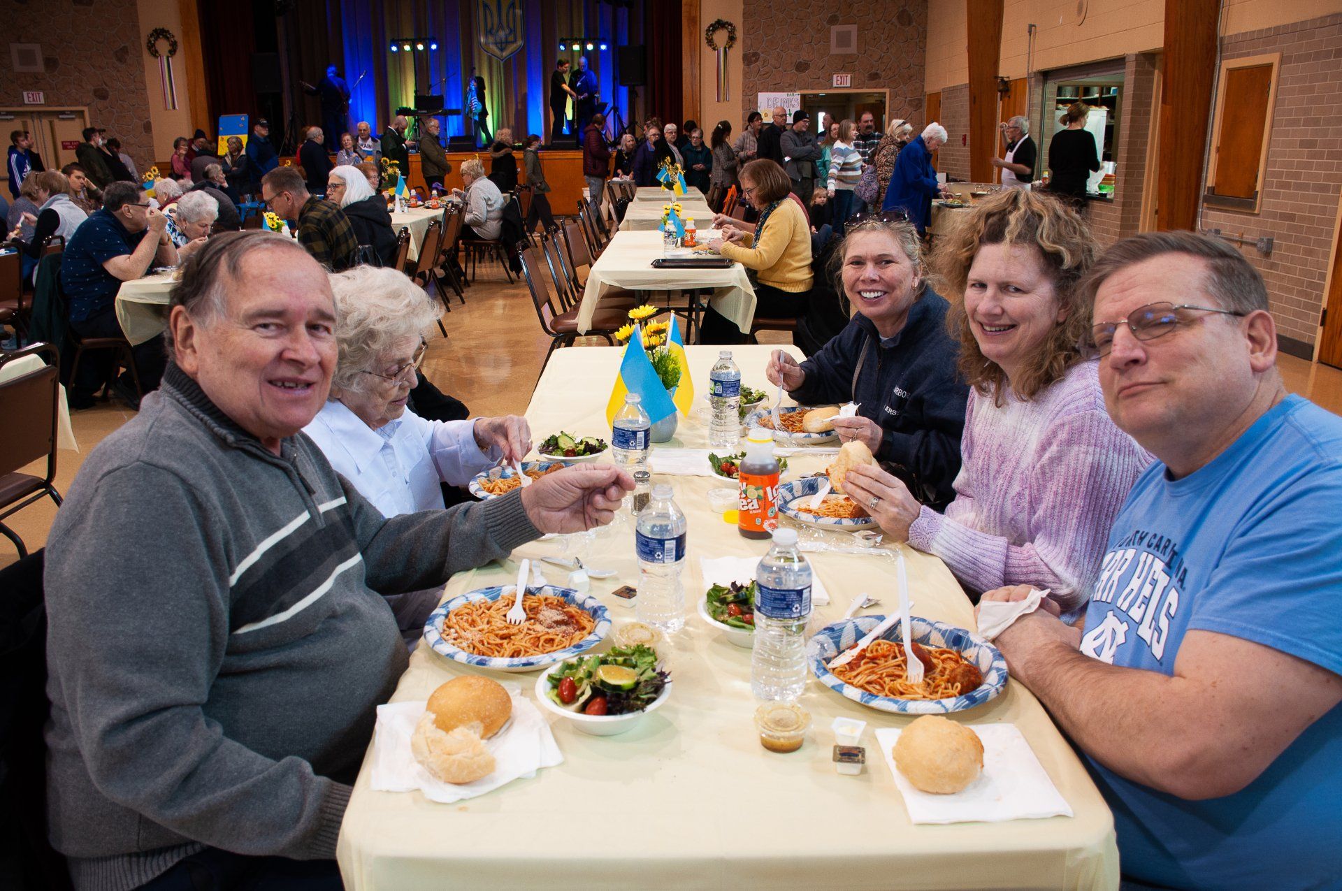 People seated at a banquet table in a hall, smiling and sharing a meal with colorful decorations.
