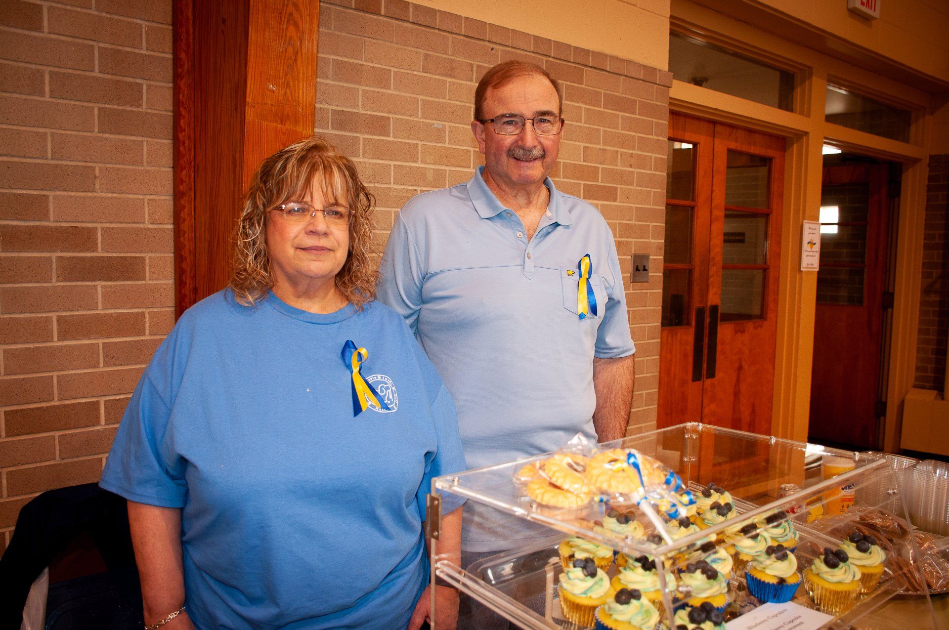 Two volunteers in blue shirts stand behind a table of baked goods in a brick hallway.