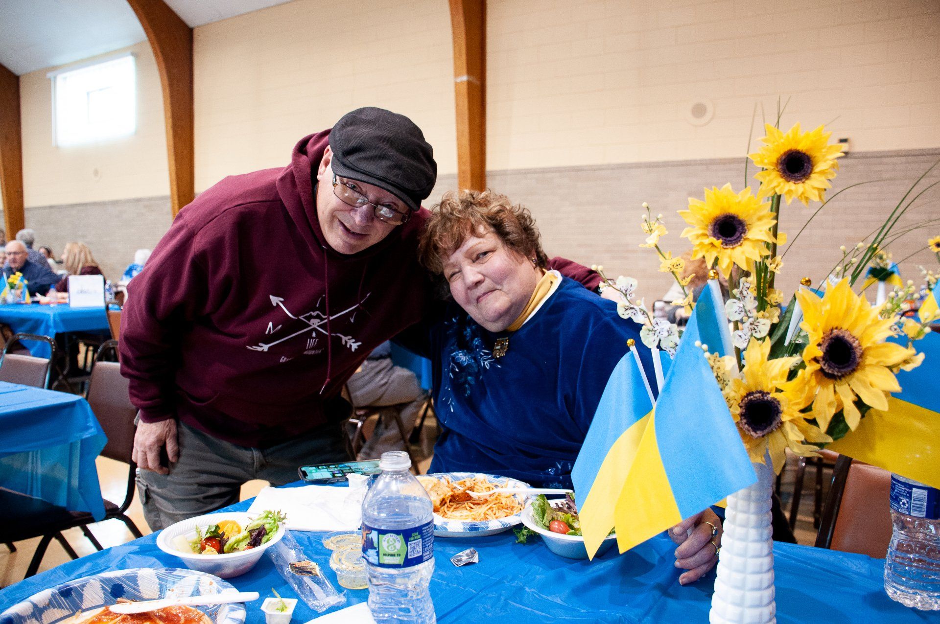 Two smiling people seated at a decorated table with sunflowers and a small blue-yellow flag.