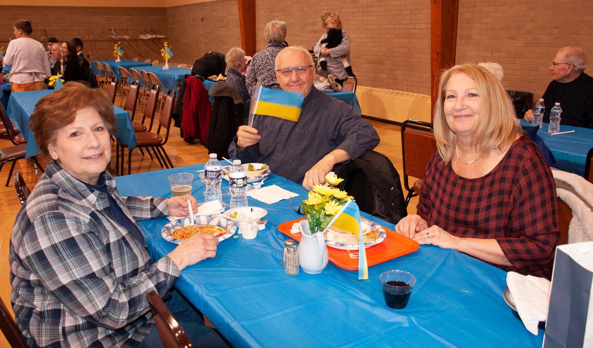 People seated at a banquet table with blue tablecloths, eating and chatting in a hall.