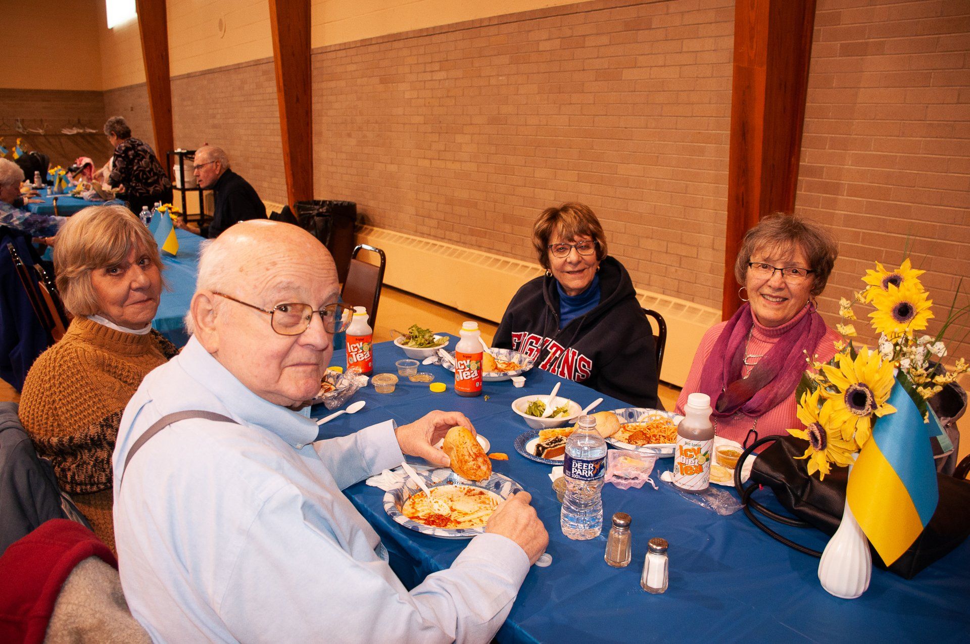 People seated at a blue table with food and sunflowers in a hall, smiling and chatting