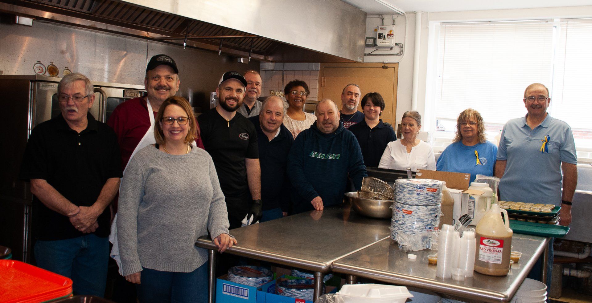 Group of people gathered in a commercial kitchen, standing behind a stainless steel prep table