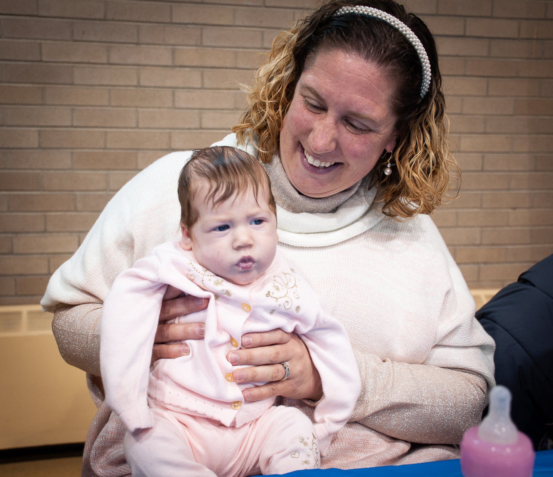 Woman holding a baby indoors, both in light pink and white clothing, smiling before a brick wall