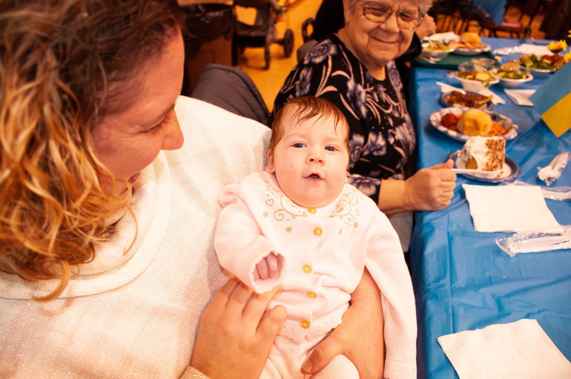 Baby in white outfit being held at a family table with food and drinks on a blue tablecloth