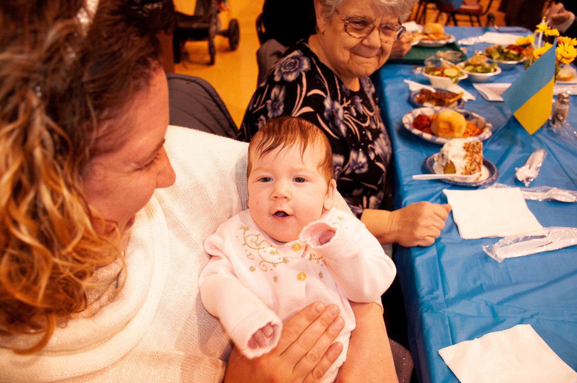 Woman holding a baby at a table during a festive meal, with an elderly woman smiling nearby