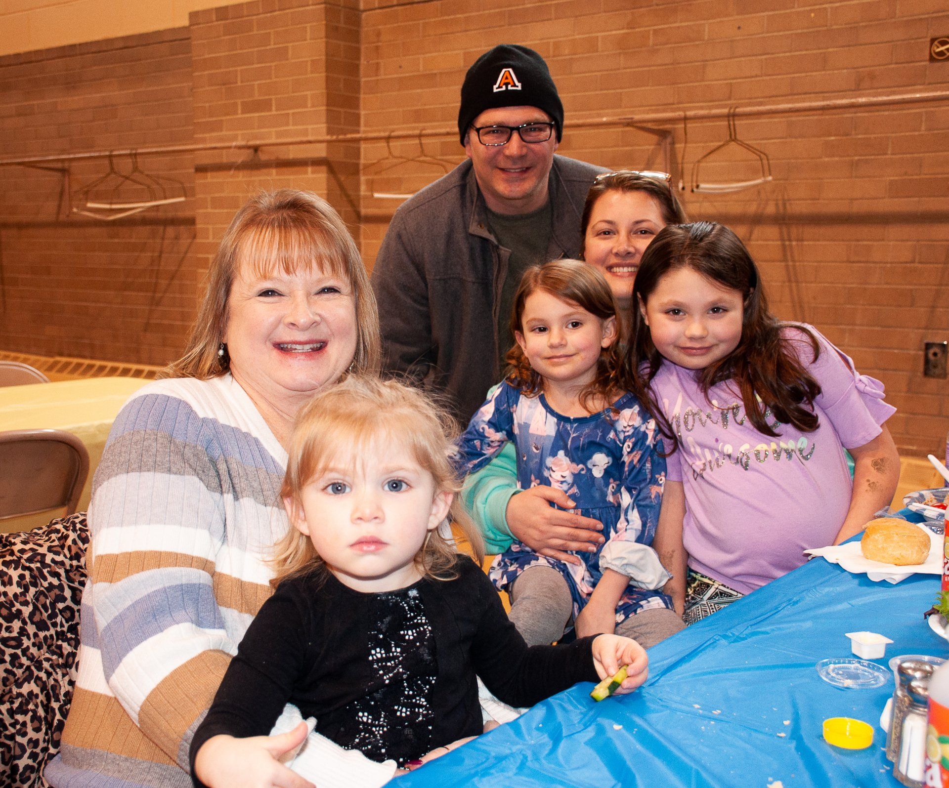 Group of people smiling at a table with a blue tablecloth at a casual indoor gathering.