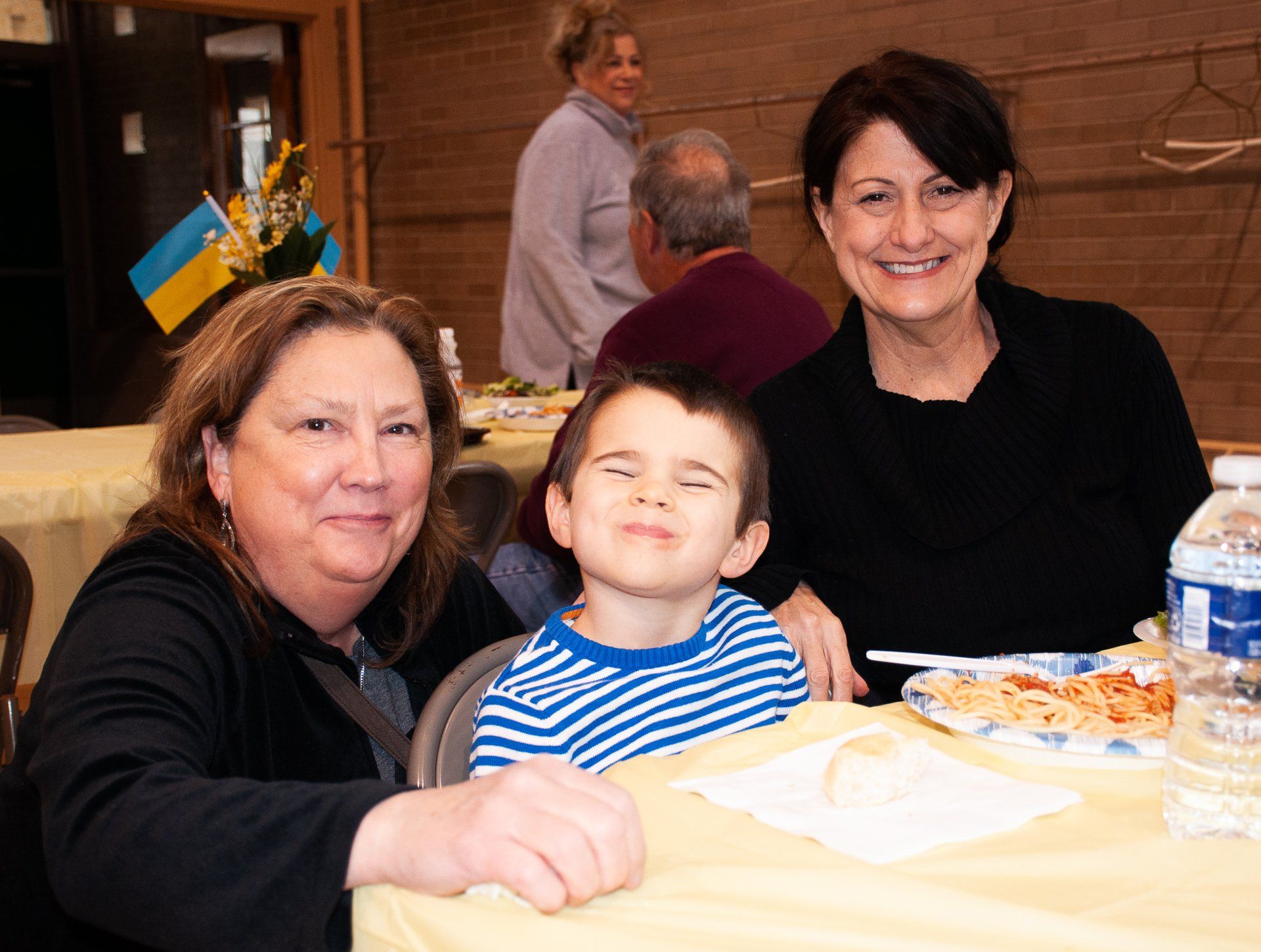 Three people sitting at a table with food and drinks, smiling indoors at a gathering.
