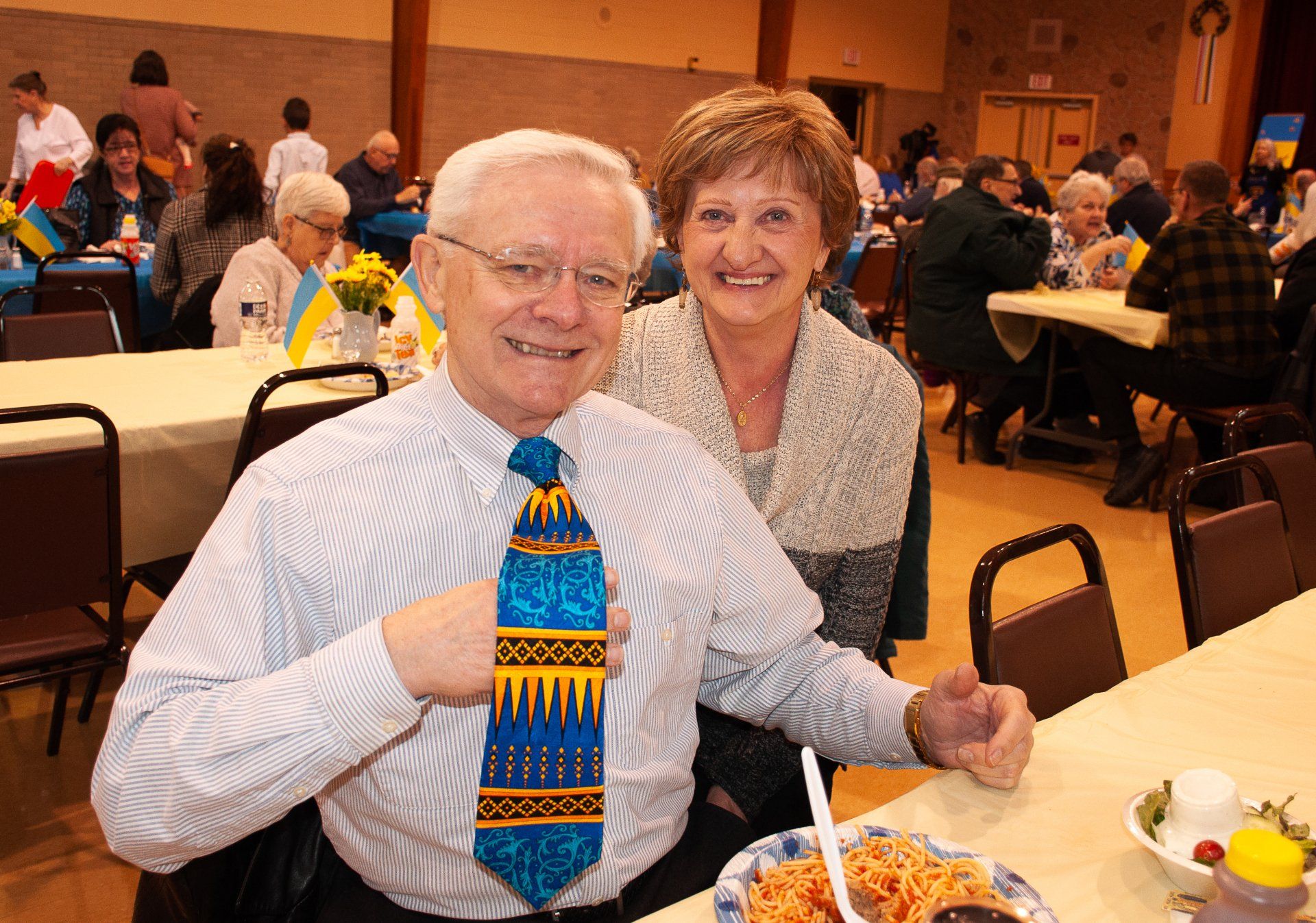 Smiling couple seated at a banquet table in a crowded hall, with plates of food and drinks nearby