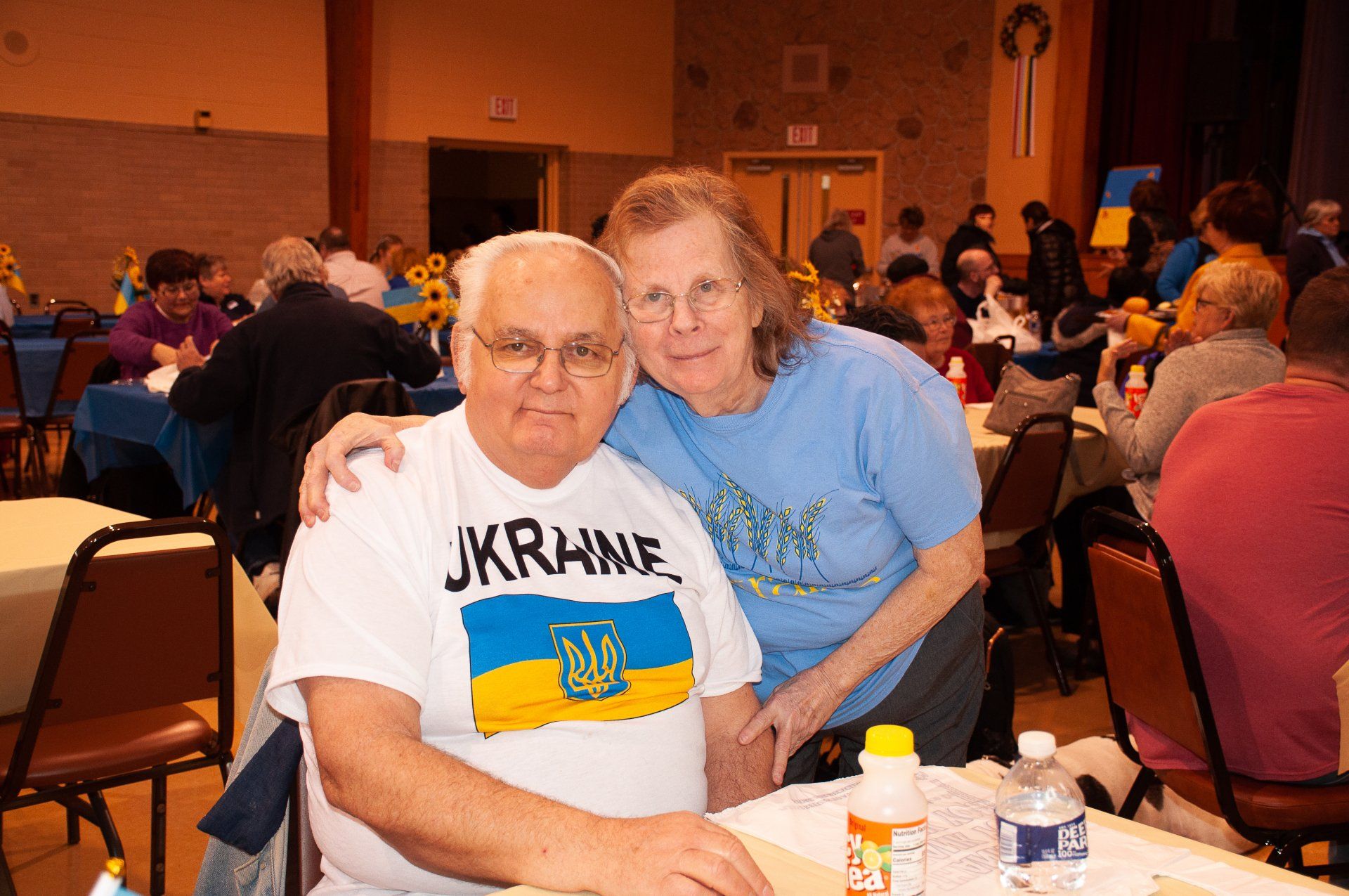 Two people seated at a banquet table in a crowded event hall, with drinks and plates on the table.