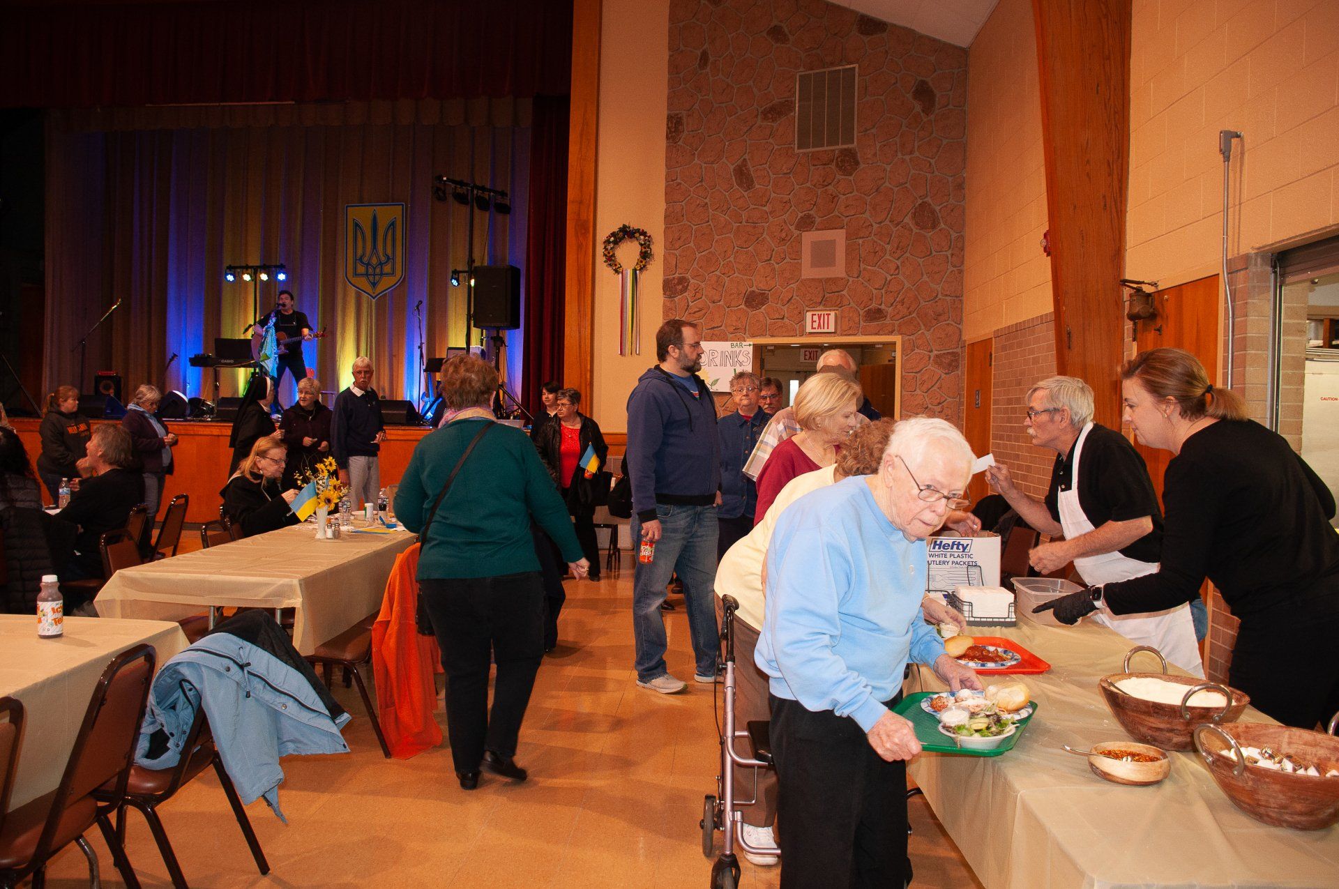 People serving food at a community banquet in a large hall with tables and stage lights