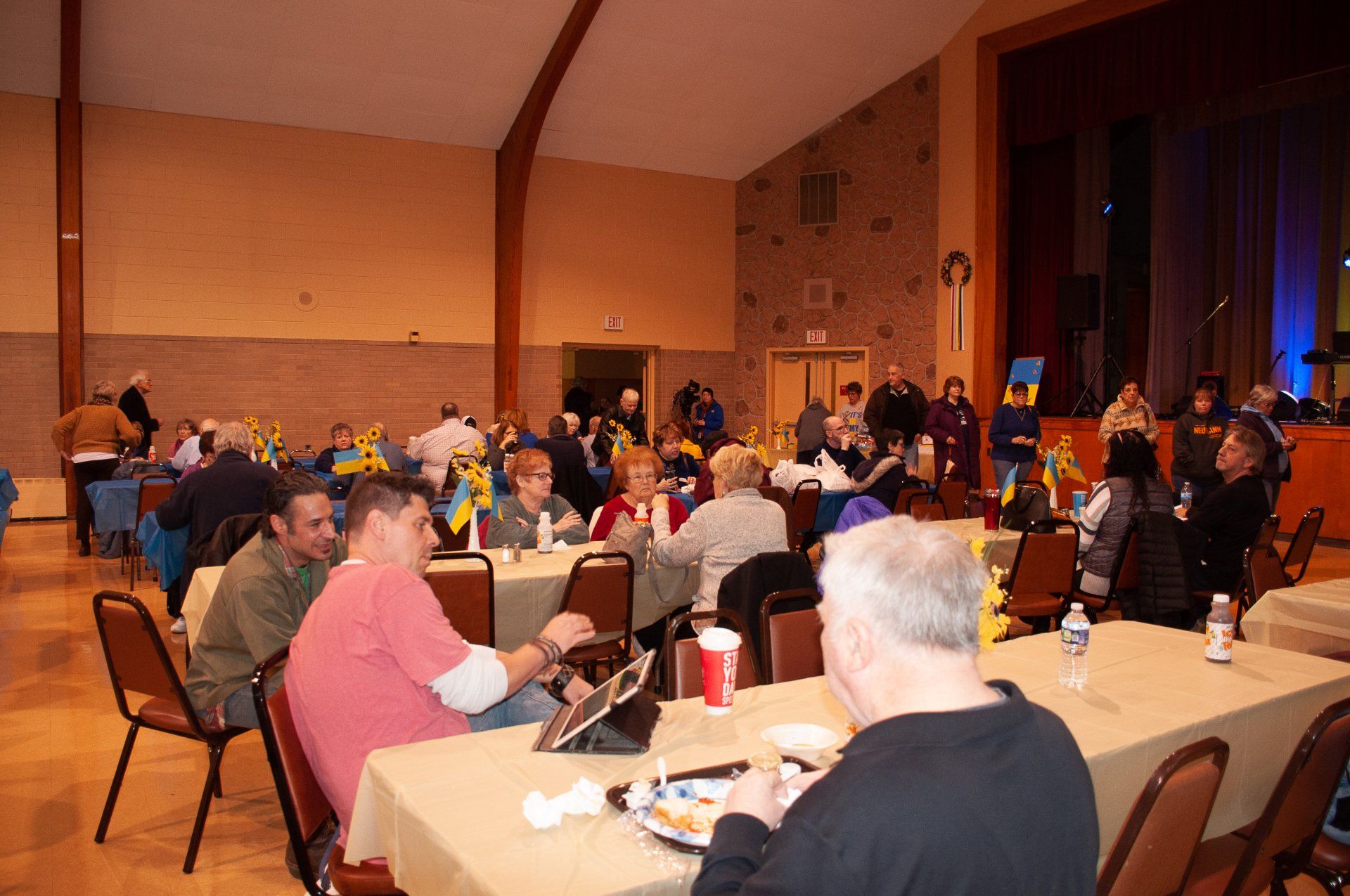 People seated at round tables in a warmly lit banquet hall, talking and eating during a gathering
