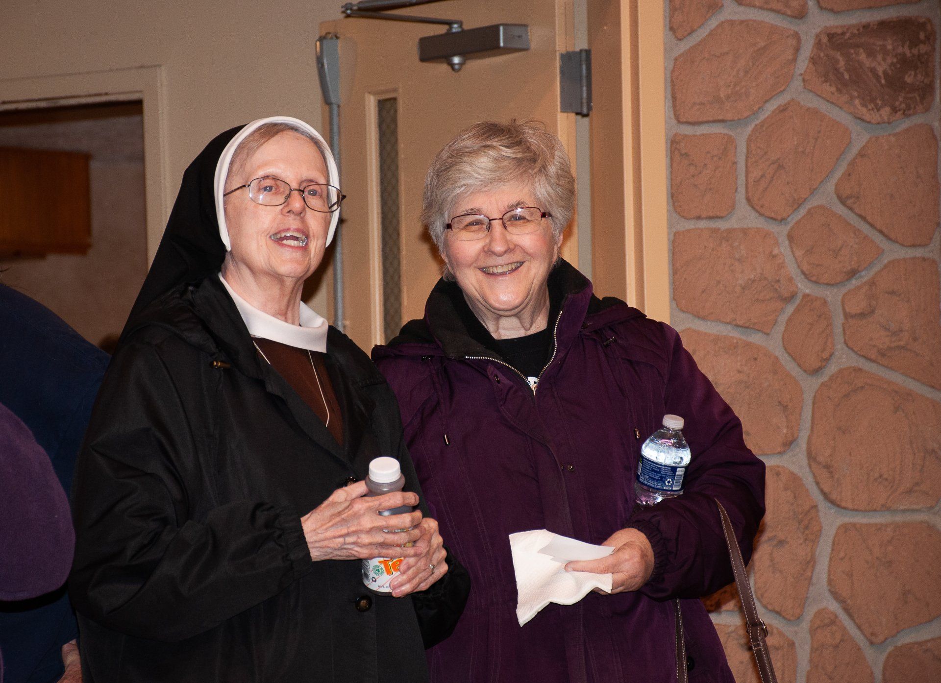 Two smiling people in coats holding water bottles and tissues indoors near a stone wall